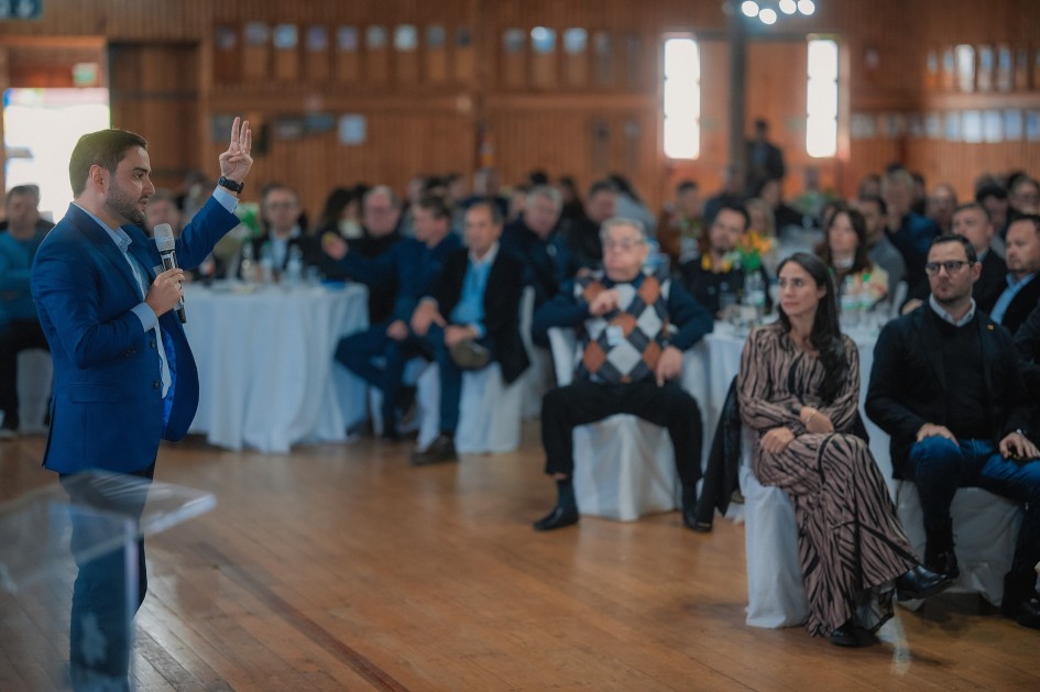 A foto mostra um ambiente interno amplo, com piso de madeira. O vice-governador Gabriel Souza, de pele clara e barba, em pé no canto esquerdo, está segurando um microfone e erguendo a mão direita, como se estivesse fazendo um ponto ou chamando a atenção. Ele veste um terno azul e uma camisa clara. À direita de Gabriel Souza, e estendendo-se ao fundo da imagem, há uma plateia diversa sentada em mesas redondas cobertas com toalhas brancas ou em cadeiras. Muitos dos rostos estão embaçados. O ambiente tem paredes de madeira e janelas altas no fundo que emitem uma luz suave.