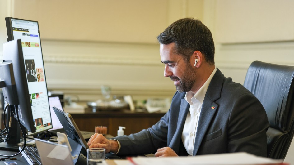 A imagem mostra um homem de meia-idade, branco, com barba curta e escura, vestindo um terno cinza-escuro e uma camisa branca aberta no colarinho. Ele está sentado em uma cadeira de escritório preta de couro, em frente a uma mesa de madeira. Em sua orelha direita, há um fone de ouvido branco sem fio.
O homem está sorrindo e olhando para baixo, para um tablet preto que está sobre a mesa. Sua mão direita está sobre a tela do tablet, como se estivesse interagindo com ele.
À esquerda do homem, há um monitor de computador vertical que exibe uma página da web com várias fotos e informações. Ao fundo, pode-se ver um escritório com paredes claras e molduras decorativas. Há também alguns objetos sobre a mesa, como um copo com um pouco de líquido e alguns papéis empilhados. A iluminação geral do ambiente é clara e natural.