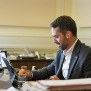 A imagem mostra um homem de meia-idade, branco, com barba curta e escura, vestindo um terno cinza-escuro e uma camisa branca aberta no colarinho. Ele está sentado em uma cadeira de escritório preta de couro, em frente a uma mesa de madeira. Em sua orelha direita, há um fone de ouvido branco sem fio.

O homem está sorrindo e olhando para baixo, para um tablet preto que está sobre a mesa. Sua mão direita está sobre a tela do tablet, como se estivesse interagindo com ele.

À esquerda do homem, há um monitor de computador vertical que exibe uma página da web com várias fotos e informações. Ao fundo, pode-se ver um escritório com paredes claras e molduras decorativas. Há também alguns objetos sobre a mesa, como um copo com um pouco de líquido e alguns papéis empilhados. A iluminação geral do ambiente é clara e natural.