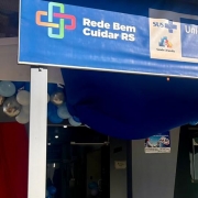 A foto exibe a fachada da Unidade Básica de Saúde em Capão do Cipó, reformada e pertencente à Rede Bem Cuidar e à Estratégia de Saúde da Família. Acima da entrada, uma grande faixa azul e branca se estende. O lado esquerdo da faixa apresenta um logotipo verde e azul com uma cruz e um coração, e o texto "Rede Bem Cuidar RS". O lado direito da faixa exibe o logotipo do SUS e "Unidade Básica de Saúde Capão do Cipó", seguido por "Secretaria Municipal de Saúde". Abaixo da faixa, à direita da entrada, há um painel verde com os dizeres "OBRA DO GOVERNO DO ESTADO" em letras brancas, e "REFORMA DE UNIDADE BÁSICA DE SAÚDE". À esquerda da entrada, balões azuis e brancos adornam o local, e um pequeno cartaz é visível em uma parede. A construção é de cor clara, com detalhes em azul. O dia está ensolarado.