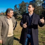 Foto dos dois homens estão de pé, conversando em um terreno aberto com grama seca. Adiló é mais velho, veste blazer bege e calça verde-oliva. Fabricio é mais jovem, usa sobretudo escuro sobre uma camisa branca e suéter azul-marinho. Ao fundo, há árvores, algumas casas e prédios. O céu está claro e azul.
