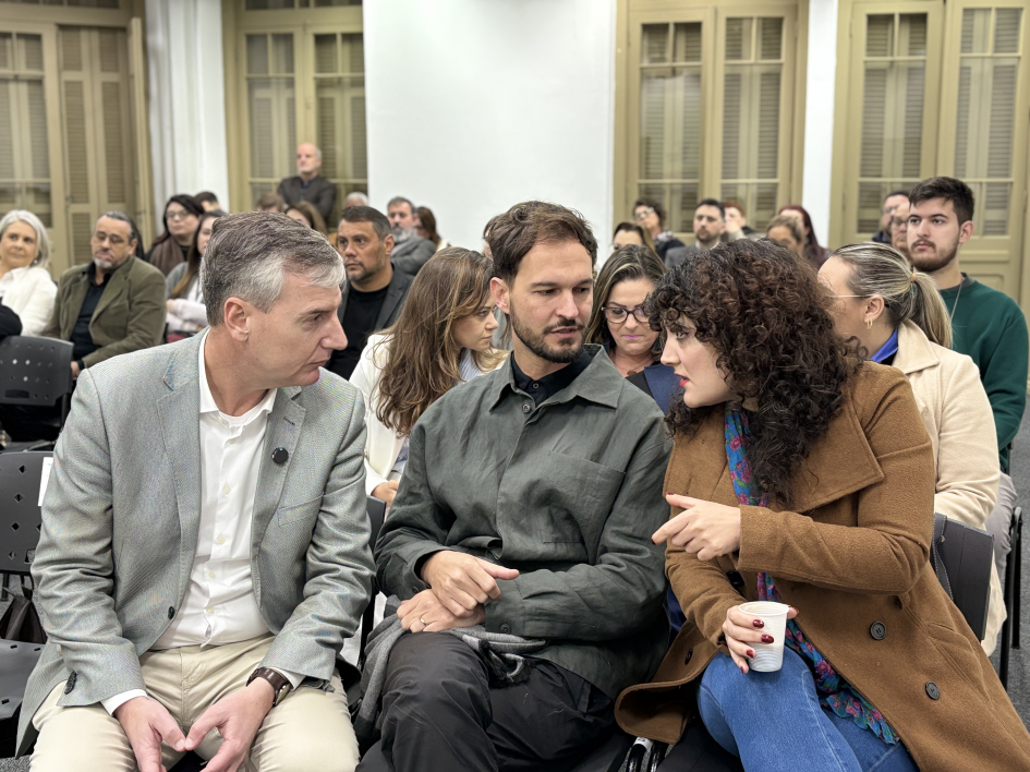 A foto em ambiente interno captura três pessoas em destaque sentadas na primeira fila de um auditório, interagindo entre si, com uma plateia ao fundo. Da esquerda para a direita, estão o secretário Eduardo Loureiro. Ele é um homem de meia-idade, cabelos grisalhos, vestindo um blazer claro e calças beges. Ele está virado para a direita, olhando e conversando com duas pessoas. Ao centro, está o diretor Rafael Balle: um homem jovem, com barba e cabelos escuros, vestindo uma camisa de tom escuro. Ele está olhando para Mariana, com as mãos cruzadas no colo. Mariana Martinez está à direita da foto. Ela é uma mulher com cabelos cacheados volumosos e escuros, vestindo um casaco marrom e jeans, com um lenço estampado azul e vermelho. Ela está virada para a esquerda, conversando e gesticulando com a mão direita, segurando um copo branco. Atrás deles, outras pessoas da plateia estão sentadas, algumas olhando para frente e outras conversando. Ao fundo, uma parede com janelas de madeira com venezianas está visível. A iluminação é suave e uniforme.