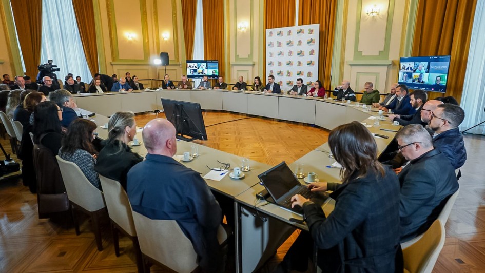 Foto de reunião formal em uma sala ampla e bem iluminada, com paredes claras, cortinas douradas e piso de madeira. Pessoas estão sentadas em torno de uma mesa grande em formato de U, todas voltadas para o centro, onde há dois monitores. Algumas pessoas usam laptops ou fazem anotações. Há câmeras e painéis com logomarcas ao fundo, além de duas telas exibindo participantes em videoconferência.