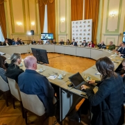 Foto de reunião formal em uma sala ampla e bem iluminada, com paredes claras, cortinas douradas e piso de madeira. Pessoas estão sentadas em torno de uma mesa grande em formato de U, todas voltadas para o centro, onde há dois monitores. Algumas pessoas usam laptops ou fazem anotações. Há câmeras e painéis com logomarcas ao fundo, além de duas telas exibindo participantes em videoconferência.
