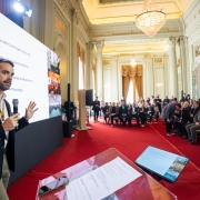 Cena em um salão amplo e sofisticado, com cortinas douradas pendendo sobre grandes janelas ao fundo e um lustre elegante no teto. À frente, o governador Eduardo Leite em pé segurando um microfone, gesticulando com uma das mãos enquanto fala. Ele está fazendo a apresentação diante de um público sentado em cadeiras enfileiradas. À sua esquerda, uma grande tela exibe uma lista de tópicos relacionados à administração pública e sustentabilidade climática.