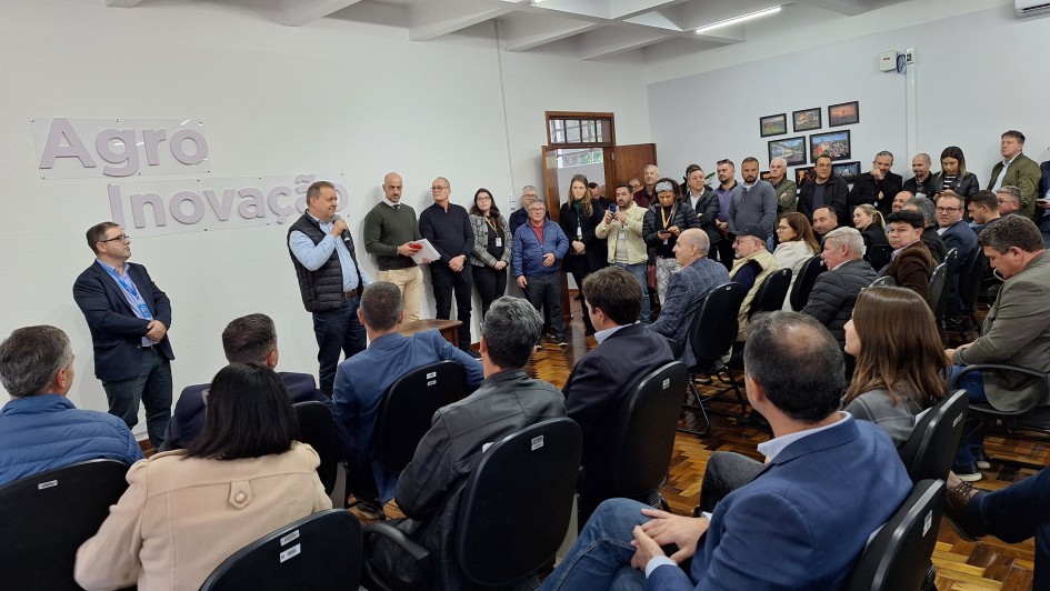A foto mostra um evento em ambiente interno com um grande grupo de pessoas. No lado esquerdo, um palco ou área elevada abriga vários homens e algumas mulheres em pé, com um dos homens no centro falando ao microfone, vestindo um colete azul e voltado para a sala. Atrás deles, na parede branca, as palavras "Agro Inovação" em relevo roxo são visíveis. A maior parte da sala é preenchida por uma plateia sentada em cadeiras escuras, virada para o palco, com muitos de costas para a câmera. Alguns quadros e uma porta de madeira são visíveis na parede à direita. A iluminação é uniforme, vinda do teto. 