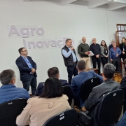 A foto mostra um evento em ambiente interno com um grande grupo de pessoas. No lado esquerdo, um palco ou área elevada abriga vários homens e algumas mulheres em pé, com um dos homens no centro falando ao microfone, vestindo um colete azul e voltado para a sala. Atrás deles, na parede branca, as palavras "Agro Inovação" em relevo roxo são visíveis. A maior parte da sala é preenchida por uma plateia sentada em cadeiras escuras, virada para o palco, com muitos de costas para a câmera. Alguns quadros e uma porta de madeira são visíveis na parede à direita. A iluminação é uniforme, vinda do teto. 