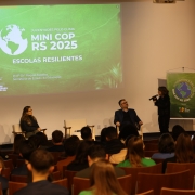 A foto mostra um evento sendo realizado em um auditório. No palco, à esquerda, dois jovens estudantes, um homem e uma mulher, ambos vestindo camisetas verdes, estão de pé ao lado de um púlpito de madeira claro. O homem está falando ao microfone. No centro do palco, duas pessoas sentam em poltronas brancas, uma mulher de óculos e um homem de blazer escuro, ambos olhando para frente. Mais à direita, outra mulher de pé, vestindo preto, segura um microfone e interage com o público ou com alguém fora do quadro. No fundo, um grande telão projeta um slide verde com a escrita "JUVENTUDES PELO CLIMA MINI COP RS 2025" e "ESCOLAS RESILIENTES", junto a logos do "GOVERNO DO ESTADO RIO GRANDE DO SUL" e "SECRETARIA DE ESTADO DA EDUCAÇÃO". À direita do palco, um banner verde com a mesma temática está visível. As primeiras filas do auditório estão cheias de estudantes, visíveis da parte de trás da cabeça, sentados em cadeiras de madeira. A iluminação é focada no palco.