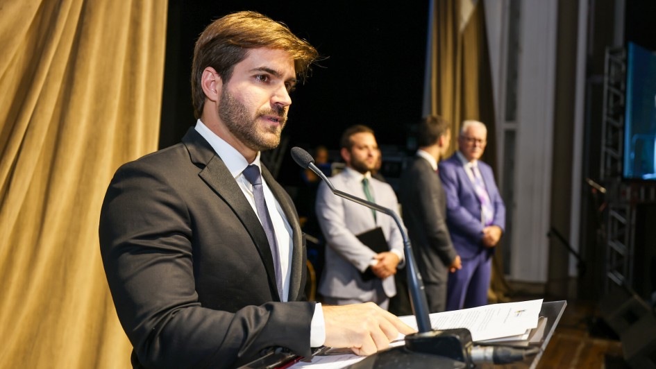 A foto mostra o médico Thalis Bolzan, um homem de pele clara e barba, com cabelos castanhos claros e bem arrumados, falando em um púlpito transparente com um microfone. Ele está ligeiramente virado para a direita e suas mãos estão sobre alguns papéis no púlpito. Ele veste um terno preto, camisa branca e gravata azul-escura. Atrás dele, no segundo plano, há três outros homens em pé, visivelmente um pouco desfocados. Ao fundo, cortinas marrons e uma tela escura à direita são visíveis.