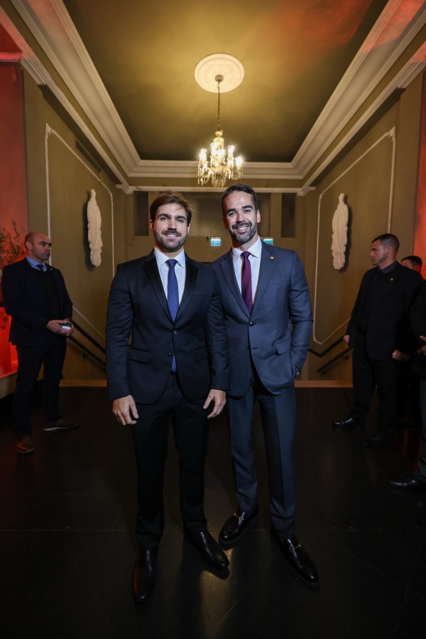 A foto mostra o governador Eduardo Leite e o médico Thalis Bolzan posando para uma fotografia em um corredor com arquitetura clássica. Eduardo Leite, com cabelo e barba castanho, veste um terno azul-escuro com camisa branca e gravata vinho, e está com as mãos nos bolsos da calça. Ao lado dele, Thalis Bolzan, com barba e cabelos castanhos claros, veste um terno preto com camisa branca e gravata azul. Ambos sorriem para a câmera, com uma postura confiante. Ao fundo, um corredor com paredes claras e molduras no teto, onde se destaca um lustre. Estátuas brancas em nichos nas paredes são visíveis. Há também algumas pessoas desfocadas em segundo plano, incluindo homens de terno.