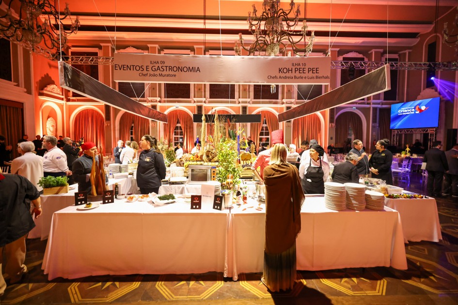 A foto mostra um amplo salão de eventos, com um buffet de comida bem iluminado no centro e muitas pessoas circulando e servindo-se. O teto do salão é alto e apresenta lustres grandes. Pendurados acima do buffet, banners brancos com a inscrição "ILHA GASTRONOMIA" são visíveis. À direita, um telão grande exibe a logomarca do "Instituto da Criança com Diabetes" com a palavra "criança" em azul e o símbolo de um coração. As paredes do salão são em tons de laranja e marrom, com detalhes em arco. Mesas com toalhas brancas e pilhas de pratos limpos estão visíveis em primeiro plano, indicando um jantar beneficente.