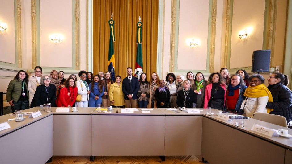 A imagem mostra um grande grupo de pessoas, predominantemente mulheres, de pé atrás de uma longa mesa de reunião em formato de "U". No centro do grupo, em pé atrás da mesa, estão o governador do Rio Grande do Sul, Eduardo Leite, e a Ministra da Mulher, ambos vestidos formalmente. Eles estão cercados por diversas mulheres, algumas sorrindo e olhando para a câmera. Ao fundo, a sala possui uma decoração clássica com paredes claras, cortinas douradas e verdes, e duas bandeiras em hastes (uma do Brasil e outra do Rio Grande do Sul). O chão é de madeira com um padrão geométrico. Há alguns equipamentos de áudio visíveis nas laterais da sala.