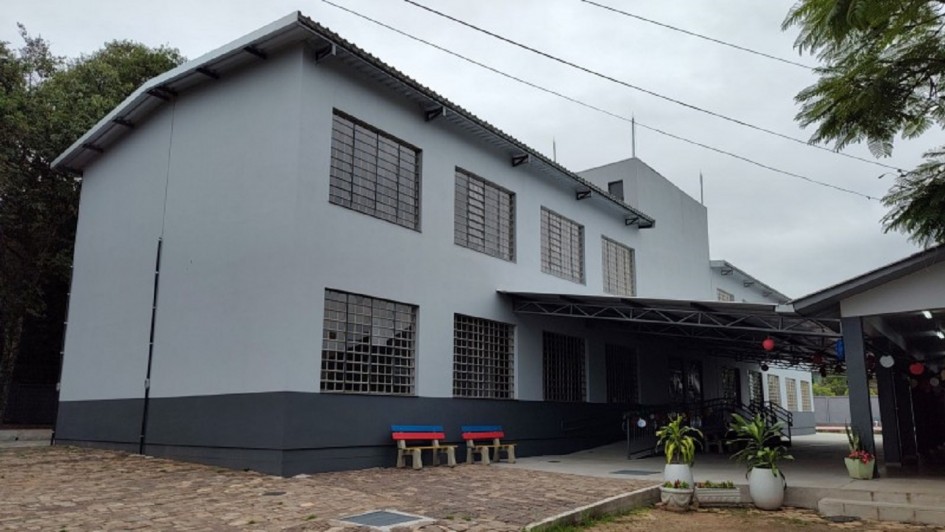 A imagem mostra a fachada de um prédio de dois andares com aparência simples e acolhedora. As paredes são cinza claro, com uma base em cinza escuro. Há grandes janelas com grades metálicas, distribuídas simetricamente. Na área da entrada, um telhado cobre a parte frontal, criando uma espécie de varanda. Em frente ao prédio, há um chão pavimentado onde se destacam dois bancos coloridos — um vermelho e outro azul — que dão um toque alegre ao ambiente.