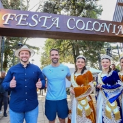 A fotografia mostra cinco pessoas (o governador Eduado Leite, o prefeito Gilberto Cezar e três mulheres da corte, posando sob um arco de madeira decorativo. Esse arco tem palavras em destaque: no topo está escrito "Festa Colonial" e na lateral esquerda, "Canela", indicando que o local é uma festa tradicional na cidade de Canela.