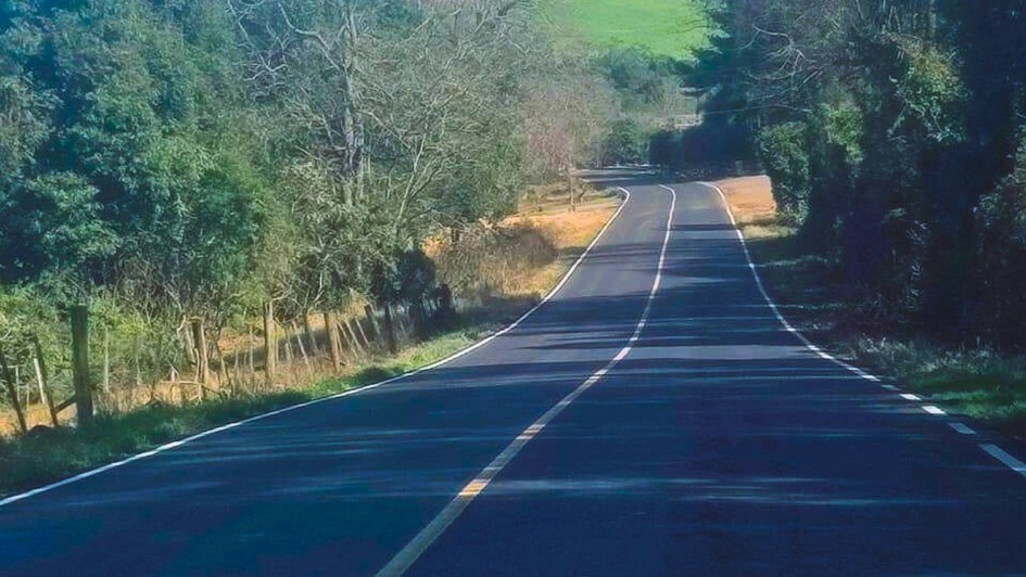 A foto mostra a estrada ERS-432, que se estende suavemente em uma curva à esquerda através de uma paisagem rural. A pista de asfalto é escura, com faixas brancas bem definidas nas laterais e uma faixa amarela central. A estrada é ladeada por árvores e vegetação densa, com folhagens verdes escuras. O sol incide diretamente sobre a via, criando um jogo de luz e sombra, e o céu azul claro é visível em pequena parte no canto superior direito. Não há veículos ou pessoas visíveis na imagem.
