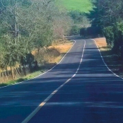 A foto mostra a estrada ERS-432, que se estende suavemente em uma curva à esquerda através de uma paisagem rural. A pista de asfalto é escura, com faixas brancas bem definidas nas laterais e uma faixa amarela central. A estrada é ladeada por árvores e vegetação densa, com folhagens verdes escuras. O sol incide diretamente sobre a via, criando um jogo de luz e sombra, e o céu azul claro é visível em pequena parte no canto superior direito. Não há veículos ou pessoas visíveis na imagem.
