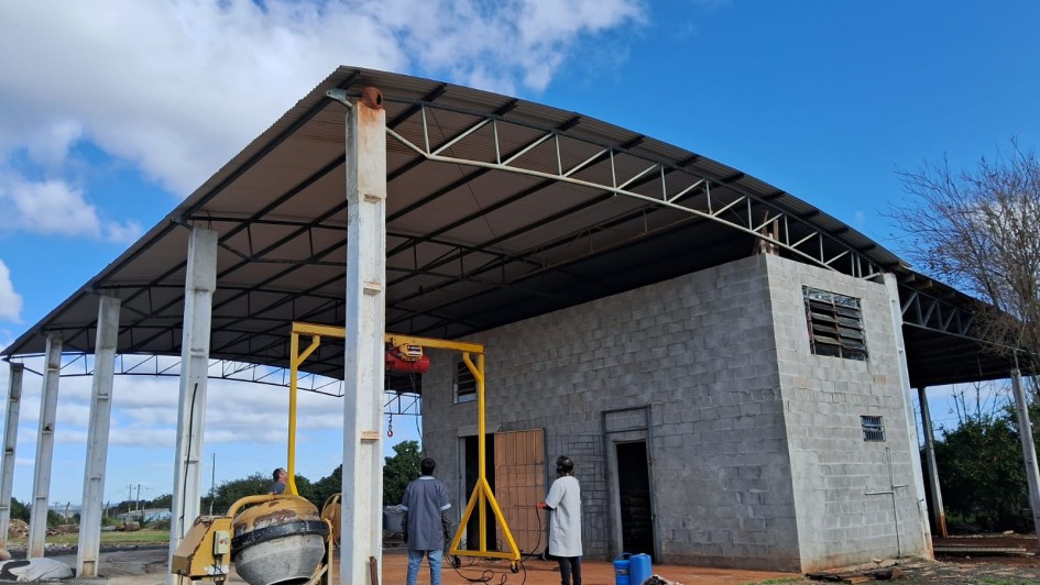 Uma imagem mostra uma construção em andamento sob um céu azul claro. No centro, há um galpão com estrutura metálica aparente e telhado, e uma parte da construção já tem paredes de blocos de concreto cinza. À esquerda, um misturador de cimento (betoneira) está estacionado. Duas pessoas, vestindo roupas de trabalho, estão em pé próximo a uma estrutura amarela tipo guindaste ou pórtico, que parece ser usada para elevação de materiais. O chão ao redor é de terra e cascalho.