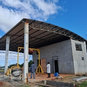 Uma imagem mostra uma construção em andamento sob um céu azul claro. No centro, há um galpão com estrutura metálica aparente e telhado, e uma parte da construção já tem paredes de blocos de concreto cinza. À esquerda, um misturador de cimento (betoneira) está estacionado. Duas pessoas, vestindo roupas de trabalho, estão em pé próximo a uma estrutura amarela tipo guindaste ou pórtico, que parece ser usada para elevação de materiais. O chão ao redor é de terra e cascalho.