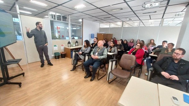 A foto mostra uma sala de treinamento ou aula com um grupo de aproximadamente vinte pessoas. Um homem com barba e camisa escura está em pé à esquerda, gesticulando e aparentemente conduzindo uma apresentação ou palestra. À sua esquerda, há uma tela grande montada em um suporte, mostrando algo fora de foco. A plateia está sentada em cadeiras de escritório, olhando para o apresentador. As pessoas são de diversas idades e vestem roupas casuais. A sala tem paredes claras com detalhes em madeira, um chão de laminado claro e iluminação fluorescente no teto. Há também janelas que parecem dar para outros espaços de escritório. Algumas mesas e objetos como garrafas de água estão visíveis na sala. O ambiente sugere um aprendizado ou uma reunião de trabalho.