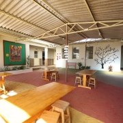 A foto mostra um pátio escolar amplo e iluminado, com teto de telhas e estrutura metálica exposta que proporciona sombra. No centro, mesas e bancos de madeira em diferentes tamanhos, sugerindo espaços para refeições ou estudo. O piso é decorado com padrões geométricos em tons de vermelho e amarelo. Ao fundo, paredes claras exibem um painel verde com adornos e cartazes coloridos, além de uma grande árvore escura pintada em uma parede branca, adicionando um toque artístico ao ambiente. Há também janelas e portas que indicam acesso a outras áreas da escola, e algumas plantas em vasos, criando um espaço acolhedor e funcional.