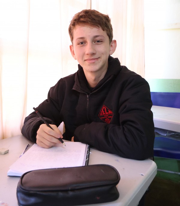 A foto mostra o aluno Júlio Cesar Medeiros. Ele é um adolescente jovem, com cabelos castanhos claros e um sorriso amigável, sentado em uma carteira escolar individual de cor clara. Ele está concentrado e segura uma caneta, com um caderno aberto à sua frente, sugerindo que está estudando ou fazendo anotações. Sobre a carteira, além do caderno e da caneta, há um estojo preto. Ele veste uma jaqueta preta com um emblema no lado esquerdo do peito. Ao fundo, uma janela com cortinas brancas translúcidas permite a entrada de luz natural, iluminando suavemente o ambiente. À direita, é possível ver parte de outra carteira escolar e uma parede de tom verde claro com um detalhe decorativo acima, que parece ser um mural.