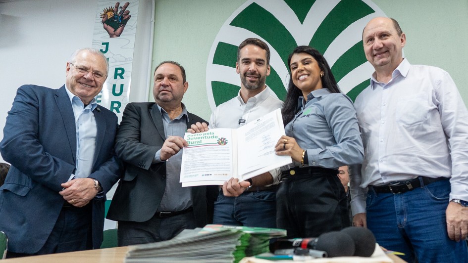 Esta é uma fotografia colorida de cinco pessoas adultas, quatro homens e uma mulher, posando lado a lado em um evento formal de assinatura de documentos.

Da esquerda para a direita:

Homem idoso, de óculos: Veste um terno azul-marinho com camisa branca e gravata escura. Ele sorri para a câmera.

Homem de meia-idade: Veste um terno escuro com camisa clara. Ele está segurando uma folha de papel branca com as duas mãos, que parece ser um documento ou um certificado, e olha para a frente.

Homem de meia-idade no centro: Veste uma camisa social clara de mangas compridas e calça escura. Ele também segura o mesmo documento que o homem à sua esquerda e sorri para a câmera. Há um logotipo com a frase "Jovens Pelo Futuro da Agricultura Rural" no documento.

Mulher jovem: Veste uma camisa cinza-claro de mangas compridas e calça escura. Ela sorri amplamente e segura a outra extremidade do documento, que parece estar aberto em uma pasta ou caderno.

Homem idoso, à direita: Veste uma camisa social branca de mangas compridas e calça jeans. Ele sorri e olha para a câmera.

Ao fundo, há uma parede verde clara com um grande logotipo estilizado em verde escuro, que se assemelha a um círculo com elementos pontiagudos. Há também um banner vertical com a letra "JUR" e uma mão estilizada segurando algo.

Na frente dos indivíduos, sobre uma mesa de madeira escura, há uma pilha de papéis, alguns cadernos e um microfone preto, sugerindo que se trata de uma coletiva de imprensa ou um evento de assinatura. A iluminação é boa e a imagem é nítida.