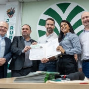 
Esta é uma fotografia colorida de cinco pessoas adultas, quatro homens e uma mulher, posando lado a lado em um evento formal de assinatura de documentos.

Da esquerda para a direita:

Homem idoso, de óculos: Veste um terno azul-marinho com camisa branca e gravata escura. Ele sorri para a câmera.

Homem de meia-idade: Veste um terno escuro com camisa clara. Ele está segurando uma folha de papel branca com as duas mãos, que parece ser um documento ou um certificado, e olha para a frente.

Homem de meia-idade no centro: Veste uma camisa social clara de mangas compridas e calça escura. Ele também segura o mesmo documento que o homem à sua esquerda e sorri para a câmera. Há um logotipo com a frase "Jovens Pelo Futuro da Agricultura Rural" no documento.

Mulher jovem: Veste uma camisa cinza-claro de mangas compridas e calça escura. Ela sorri amplamente e segura a outra extremidade do documento, que parece estar aberto em uma pasta ou caderno.

Homem idoso, à direita: Veste uma camisa social branca de mangas compridas e calça jeans. Ele sorri e olha para a câmera.

Ao fundo, há uma parede verde clara com um grande logotipo estilizado em verde escuro, que se assemelha a um círculo com elementos pontiagudos. Há também um banner vertical com a letra "JUR" e uma mão estilizada segurando algo.

Na frente dos indivíduos, sobre uma mesa de madeira escura, há uma pilha de papéis, alguns cadernos e um microfone preto, sugerindo que se trata de uma coletiva de imprensa ou um evento de assinatura. A iluminação é boa e a imagem é nítida.