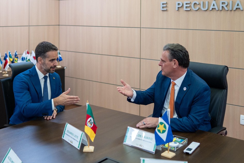 A foto mostra o governador Eduardo Leite e o ministro da Agricultura e Pecuária, Carlos Fávaro, sentados frente a frente em uma mesa de reunião espaçosa dentro de um escritório. Eduardo Leite, à esquerda, usa um terno azul escuro e gravata da mesma cor, com barba bem feita, e gesticula com a mão direita aberta em direção ao ministro. Carlos Fávaro, à direita, também veste um terno azul, mas com uma gravata laranja vibrante, e responde ao governador com uma expressão séria e gestos com as mãos. Sobre a mesa de madeira escura, entre eles, há pequenas bandeiras do Rio Grande do Sul e do Brasil, além de placas de identificação com seus nomes. Ao fundo, uma parede de madeira clara exibe parte da palavra "PECUÁRIA", sugerindo o local. O ambiente é formal e bem iluminado, indicando uma reunião de trabalho importante.