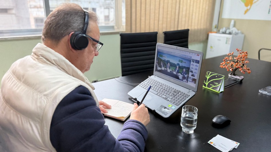 A foto mostra o Secretário da Seapi, Edivilson Brum, de costas, participando de uma reunião virtual. Ele está sentado em uma mesa preta, usando fones de ouvido pretos e óculos, e veste uma jaqueta puffer clara por cima de uma blusa escura. Em sua frente, um notebook exibe a tela da reunião com várias pessoas em vídeo, enquanto ele faz anotações em um caderno com uma caneta. Sobre a mesa, há também um copo com água, um mouse de computador e alguns papéis. Ao fundo, uma janela com persianas, uma parede branca com um mapa do estado e um arranjo de pequenas árvores alaranjadas, além de cadeiras vazias, indicam um ambiente de escritório.