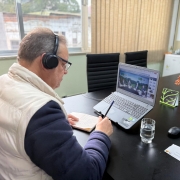 A foto mostra o Secretário da Seapi, Edivilson Brum, de costas, participando de uma reunião virtual. Ele está sentado em uma mesa preta, usando fones de ouvido pretos e óculos, e veste uma jaqueta puffer clara por cima de uma blusa escura. Em sua frente, um notebook exibe a tela da reunião com várias pessoas em vídeo, enquanto ele faz anotações em um caderno com uma caneta. Sobre a mesa, há também um copo com água, um mouse de computador e alguns papéis. Ao fundo, uma janela com persianas, uma parede branca com um mapa do estado e um arranjo de pequenas árvores alaranjadas, além de cadeiras vazias, indicam um ambiente de escritório.
