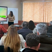 Esta é uma fotografia tirada de um ângulo posterior da plateia, mostrando uma apresentação em uma sala de aula ou auditório pequeno.

No centro da imagem, em primeiro plano, várias pessoas estão sentadas de costas para o observador, olhando para frente, onde uma mulher está apresentando. A plateia é composta principalmente por adultos e idosos.

À esquerda, no fundo, uma mulher vestindo uma blusa bege clara está em pé ao lado de uma grande tela de projeção branca. Na tela, há um slide com o título "CONSULTA POPULAR 2025/2026" e "Como participar?", com informações divididas em três etapas numeradas (01, 02, 03) e caixas de texto abaixo. A mulher aponta para a tela com a mão direita.

À direita, no fundo da sala, outra mulher está em pé, atrás de uma mesa, com alguns papéis na mão. A sala tem paredes claras e uma grande janela coberta por persianas verticais bege no lado direito. O teto é de cor clara, com luminárias embutidas. A iluminação é boa e a imagem é nítida.