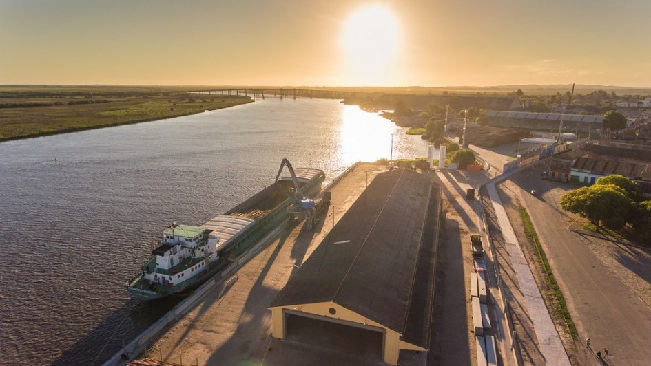 A foto mostra uma vista aérea do Porto do Rio Grande ao pôr do sol, com o sol brilhante refletindo na superfície da água do rio. No primeiro plano, à esquerda, uma barcaça alongada e de cor esverdeada está atracada no cais, recebendo carga de um braço mecânico de cor clara, provavelmente grãos. Ao lado do cais, há um longo galpão com telhado escuro e um amplo pátio ensolarado. Uma estrada margeia o cais, com alguns veículos estacionados e árvores. Do outro lado do rio, estende-se uma vasta área de vegetação e, ao fundo, uma ponte se projeta sobre as águas, conectando as margens e completando a paisagem do porto em plena atividade.