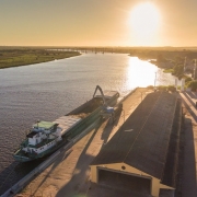 A foto mostra uma vista aérea do Porto do Rio Grande ao pôr do sol, com o sol brilhante refletindo na superfície da água do rio. No primeiro plano, à esquerda, uma barcaça alongada e de cor esverdeada está atracada no cais, recebendo carga de um braço mecânico de cor clara, provavelmente grãos. Ao lado do cais, há um longo galpão com telhado escuro e um amplo pátio ensolarado. Uma estrada margeia o cais, com alguns veículos estacionados e árvores. Do outro lado do rio, estende-se uma vasta área de vegetação e, ao fundo, uma ponte se projeta sobre as águas, conectando as margens e completando a paisagem do porto em plena atividade.