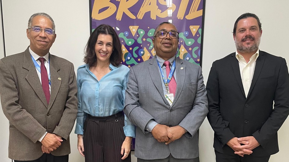 A foto mostra quatro pessoas posando para a câmera em um ambiente interno. Da esquerda para a direita, Henrique Miguel, Secretário de Ciência e Tecnologia do MCTI, sorri usando óculos e um terno marrom com gravata vinho. Ao lado dele, Simone Stülp, Secretária, sorri para a câmera vestindo uma camisa azul clara e calça escura. No centro, um homem usa óculos, terno cinza com gravata rosa e um crachá pendurado no pescoço. À direita, Murilo Pessatti, CEO da Chipus, sorri usando um blazer preto sobre uma camisa branca. Ao fundo, uma parede clara com um grande cartaz colorido com a palavra "BRASIL" escrita em letras grandes e roxas na parte superior.