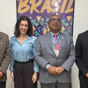 A foto mostra quatro pessoas posando para a câmera em um ambiente interno. Da esquerda para a direita, Henrique Miguel, Secretário de Ciência e Tecnologia do MCTI, sorri usando óculos e um terno marrom com gravata vinho. Ao lado dele, Simone Stülp, Secretária, sorri para a câmera vestindo uma camisa azul clara e calça escura. No centro, um homem usa óculos, terno cinza com gravata rosa e um crachá pendurado no pescoço. À direita, Murilo Pessatti, CEO da Chipus, sorri usando um blazer preto sobre uma camisa branca. Ao fundo, uma parede clara com um grande cartaz colorido com a palavra "BRASIL" escrita em letras grandes e roxas na parte superior.
