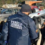 A foto mostra a parte de trás de dois homens, ambos vestindo casacos escuros com logotipos e bonés, em um ambiente externo que parece ser um desmanche ilegal de veículos. O homem da esquerda usa uma jaqueta azul escura com a inscrição "PERÍCIA CRIMINAL RIO GRANDE DO SUL" nas costas e calça jeans. O homem da direita veste uma jaqueta preta com "SECRETARIA DA SEGURANÇA PÚBLICA" nas costas e calça escura. Eles estão parados em meio a uma grande quantidade de carcaças e peças de carros amontoadas e enferrujadas, sob um céu azul e claro. Ao fundo, árvores sem folhas e algumas casas modestas completam a paisagem do local, que reflete a desordem do ambiente de apreensão.
