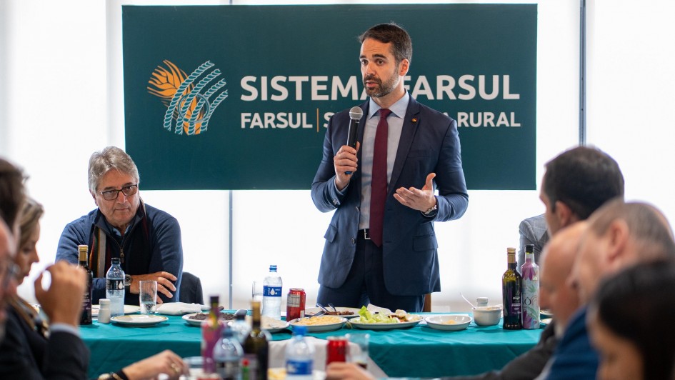 Esta é uma fotografia colorida de um evento formal, onde um homem está discursando para um grupo de pessoas sentadas ao redor de uma mesa.
No centro da imagem, em primeiro plano, um homem com barba, vestindo um terno azul-marinho, camisa branca e gravata escura, está em pé e segura um microfone com a mão direita, enquanto gesticula com a mão esquerda. Ele parece estar fazendo um discurso ou apresentação.
Atrás dele, há um grande banner azul-petróleo com o logotipo e o nome "SISTEMA FARSUL" em letras brancas, com um emblema laranja e azul de espigas de trigo ou plantas agrícolas à esquerda.
Ao redor da mesa, coberta por uma toalha azul-petróleo, várias pessoas estão sentadas, algumas comendo ou prestando atenção ao orador. No lado esquerdo, um homem de óculos, com cabelo grisalho, veste uma camisa escura com um lenço azul enrolado no pescoço. Há pratos com comida, garrafas de água e outros itens sobre a mesa.
A iluminação é boa e o ambiente parece ser interno, possivelmente um salão de eventos ou sala de reuniões.