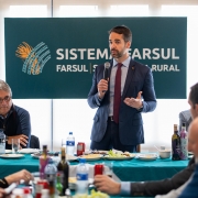 Esta é uma fotografia colorida de um evento formal, onde um homem está discursando para um grupo de pessoas sentadas ao redor de uma mesa.

No centro da imagem, em primeiro plano, um homem com barba, vestindo um terno azul-marinho, camisa branca e gravata escura, está em pé e segura um microfone com a mão direita, enquanto gesticula com a mão esquerda. Ele parece estar fazendo um discurso ou apresentação.

Atrás dele, há um grande banner azul-petróleo com o logotipo e o nome "SISTEMA FARSUL" em letras brancas, com um emblema laranja e azul de espigas de trigo ou plantas agrícolas à esquerda.

Ao redor da mesa, coberta por uma toalha azul-petróleo, várias pessoas estão sentadas, algumas comendo ou prestando atenção ao orador. No lado esquerdo, um homem de óculos, com cabelo grisalho, veste uma camisa escura com um lenço azul enrolado no pescoço. Há pratos com comida, garrafas de água e outros itens sobre a mesa.

A iluminação é boa e o ambiente parece ser interno, possivelmente um salão de eventos ou sala de reuniões.