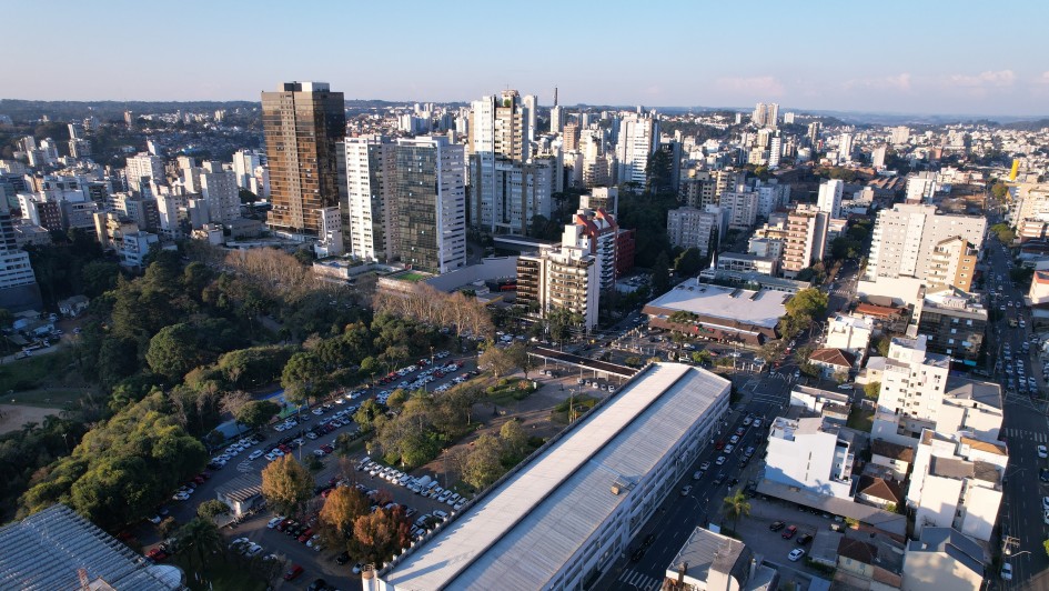 A foto mostra uma vista aérea panorâmica da cidade de Caxias do Sul durante o dia, sob um céu claro e azul. Em primeiro plano, no centro da imagem, destaca-se um longo edifício retangular com telhado claro, cercado por uma área de estacionamento com muitos carros e uma área verde arborizada à esquerda. Ao redor, a paisagem é dominada por uma densidade de edifícios de diferentes alturas e estilos, que se estendem até o horizonte. Há tanto prédios mais antigos quanto construções modernas de múltiplos andares. No canto inferior direito, uma rua movimentada com carros visíveis, sugerindo a vida urbana da cidade. A iluminação solar suave indica um fim de tarde ou começo de manhã, realçando os contornos dos edifícios e a vasta extensão urbana.