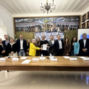 Foto posada de várias pessoas de pé atrás de uma grande mesa. Elas estão de pé sorrindo para a câmera. Ao meio, algumas delas seguram e exibem um documento.