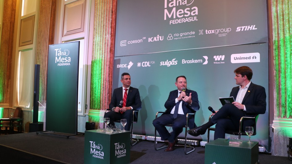 Esta é uma fotografia que mostra três homens sentados em cadeiras de palco, participando de um painel de discussão ou entrevista.

No centro da imagem, um homem de meia-idade com cabelo escuro, vestindo um terno cinza escuro, camisa branca e gravata azul, está segurando um microfone com a mão direita e gesticulando com a esquerda, enquanto fala.

À sua esquerda, outro homem de terno escuro e gravata vermelha está sentado em silêncio, observando. À sua direita, um homem mais jovem, com cabelo castanho claro e vestindo um blazer escuro sobre uma camisa branca, está sentado com as pernas cruzadas, segurando uma pequena pasta ou tablet e um microfone, olhando para o orador central.

Eles estão sentados em cadeiras estofadas escuras. No chão, em frente a cada um, há pequenos suportes verdes com copos de água e alguns itens. Há também cubos verdes com o logotipo da "FEDERASUL".

Ao fundo, há um grande painel cinza escuro com o título "Tá na Mesa FEDERASUL" em letras brancas. Abaixo, há uma série de logotipos de empresas patrocinadoras, incluindo "Corsan", "Icatu", "Rio Grande Energia", "Taxgroup", "Stihl", "Bradesco", "CDL Porto Alegre", "Sulgás", "Braskem", "Wilson Sons" e "Unimed".

O cenário tem pilares decorados em tons de dourado e verde e o teto é alto e ornamentado. Há uma iluminação focada nos participantes do painel.