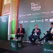 Esta é uma fotografia que mostra três homens sentados em cadeiras de palco, participando de um painel de discussão ou entrevista.

No centro da imagem, um homem de meia-idade com cabelo escuro, vestindo um terno cinza escuro, camisa branca e gravata azul, está segurando um microfone com a mão direita e gesticulando com a esquerda, enquanto fala.

À sua esquerda, outro homem de terno escuro e gravata vermelha está sentado em silêncio, observando. À sua direita, um homem mais jovem, com cabelo castanho claro e vestindo um blazer escuro sobre uma camisa branca, está sentado com as pernas cruzadas, segurando uma pequena pasta ou tablet e um microfone, olhando para o orador central.

Eles estão sentados em cadeiras estofadas escuras. No chão, em frente a cada um, há pequenos suportes verdes com copos de água e alguns itens. Há também cubos verdes com o logotipo da "FEDERASUL".

Ao fundo, há um grande painel cinza escuro com o título "Tá na Mesa FEDERASUL" em letras brancas. Abaixo, há uma série de logotipos de empresas patrocinadoras, incluindo "Corsan", "Icatu", "Rio Grande Energia", "Taxgroup", "Stihl", "Bradesco", "CDL Porto Alegre", "Sulgás", "Braskem", "Wilson Sons" e "Unimed".

O cenário tem pilares decorados em tons de dourado e verde e o teto é alto e ornamentado. Há uma iluminação focada nos participantes do painel.