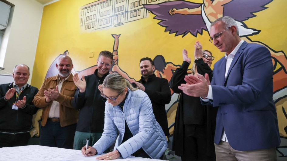Um grupo de pessoas está reunido em volta de uma mesa. No centro, alguém assina um documento enquanto os outros batem palmas, comemorando o momento. Ao fundo, há um mural colorido com um prédio e uma figura com asas, criando uma atmosfera vibrante e simbólica.