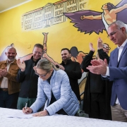Um grupo de pessoas está reunido em volta de uma mesa. No centro, alguém assina um documento enquanto os outros batem palmas, comemorando o momento. Ao fundo, há um mural colorido com um prédio e uma figura com asas, criando uma atmosfera vibrante e simbólica.