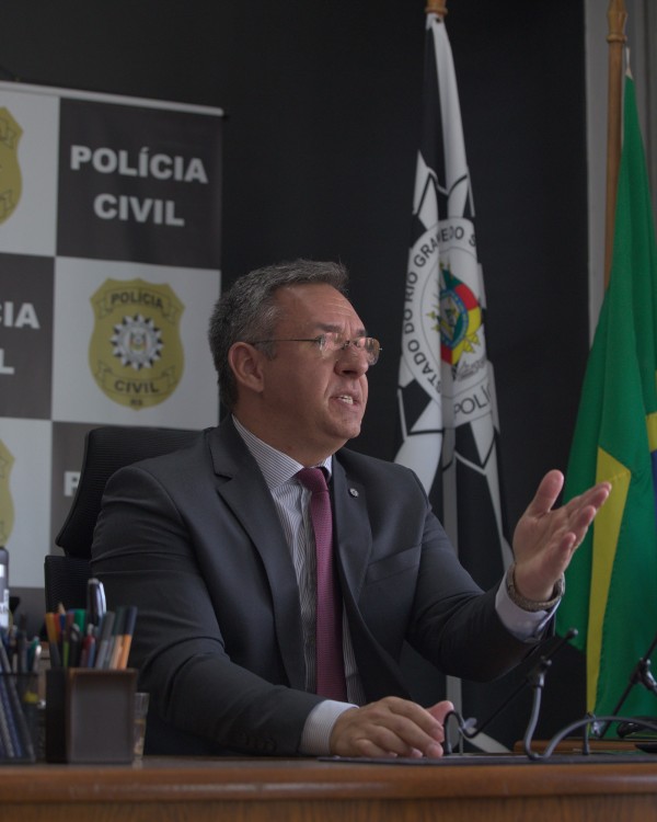 A foto mostra Augusto Cavalheiro Neto, delegado regional da Polícia Civil na Serra, sentado a uma mesa, de perfil, falando e gesticulando com a mão esquerda aberta. Ele usa um terno escuro, camisa social clara e gravata vinho listrada, com óculos de grau. A sua frente, há um organizador de canetas e objetos de escritório. Ao fundo, um banner com o brasão da Polícia Civil e as palavras "POLÍCIA CIVIL" é visível, parcialmente oculto por uma bandeira do Rio Grande do Sul e uma bandeira do Brasil. O ambiente parece ser um escritório ou sala de coletivas, indicando uma comunicação oficial ou declaração.