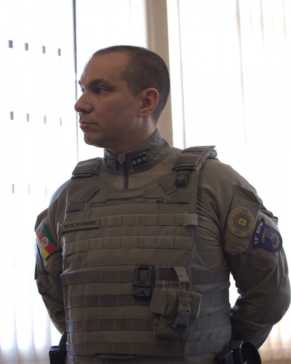 A foto mostra o Tenente-coronel Márcio Borba Fernandes, comandante do 12º BPM, em pose séria e de perfil, olhando para a esquerda da imagem. Ele veste um uniforme militar tático na cor cáqui, incluindo um colete balístico com bolsos e presilhas. Nas ombreiras do colete, há distintivos com três estrelas, e no ombro esquerdo, a bandeira do Rio Grande do Sul está visível. No braço direito, um emblema azul com a inscrição "12º BPM". Ele tem cabelo curto e escuro. Ao fundo, uma janela com persianas verticais claras e um ambiente interno bem iluminado.