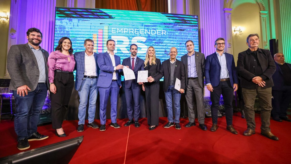 Nove pessoas estão em pé sobre um tapete vermelho, em frente a uma tela grande que exibe a frase EMPREENDER RS. Algumas no centro estão segurando documentos. A imagem transmite um momento de celebração formal.