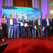 Nove pessoas estão em pé sobre um tapete vermelho, em frente a uma tela grande que exibe a frase EMPREENDER RS. Algumas no centro estão segurando documentos. A imagem transmite um momento de celebração formal.