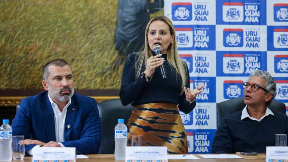 Esta é uma fotografia de três pessoas, uma mulher e dois homens, sentadas em uma mesa em um evento, com a mulher no centro falando ao microfone.
No centro da imagem, uma mulher com cabelos loiros longos, vestindo uma blusa preta de manga comprida e uma saia com estampa abstrata em tons de marrom e bege, está de pé e gesticulando com as mãos enquanto fala em um microfone preto. Ela usa um anel grande e chamativo na mão direita.
À esquerda da mulher, um homem com barba grisalha e cabelos curtos, vestindo um blazer azul-marinho sobre uma camisa branca, está sentado à mesa olhando para a frente. À sua frente, há uma garrafa de água e uma placa de identificação com o nome "BRUNO SILVEIRA".
À direita da mulher, outro homem com cabelos grisalhos e óculos, vestindo um suéter escuro, está sentado olhando para a mulher enquanto ela fala. À sua frente, há uma placa de identificação com o nome "FREDERICO". Uma garrafa de água também está visível na mesa.
O fundo da imagem apresenta uma parede com um grande banner branco e azul que exibe repetidamente o logotipo da "PREFEITURA DE URUGUAIANA" e o nome "URUGUAIANA". À esquerda do banner, há uma pintura emoldurada na parede com detalhes dourados. A mesa é de madeira escura e a iluminação é clara e uniforme.