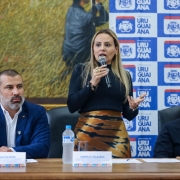 Esta é uma fotografia de três pessoas, uma mulher e dois homens, sentadas em uma mesa em um evento, com a mulher no centro falando ao microfone.

No centro da imagem, uma mulher com cabelos loiros longos, vestindo uma blusa preta de manga comprida e uma saia com estampa abstrata em tons de marrom e bege, está de pé e gesticulando com as mãos enquanto fala em um microfone preto. Ela usa um anel grande e chamativo na mão direita.

À esquerda da mulher, um homem com barba grisalha e cabelos curtos, vestindo um blazer azul-marinho sobre uma camisa branca, está sentado à mesa olhando para a frente. À sua frente, há uma garrafa de água e uma placa de identificação com o nome "BRUNO SILVEIRA".

À direita da mulher, outro homem com cabelos grisalhos e óculos, vestindo um suéter escuro, está sentado olhando para a mulher enquanto ela fala. À sua frente, há uma placa de identificação com o nome "FREDERICO". Uma garrafa de água também está visível na mesa.

O fundo da imagem apresenta uma parede com um grande banner branco e azul que exibe repetidamente o logotipo da "PREFEITURA DE URUGUAIANA" e o nome "URUGUAIANA". À esquerda do banner, há uma pintura emoldurada na parede com detalhes dourados. A mesa é de madeira escura e a iluminação é clara e uniforme.